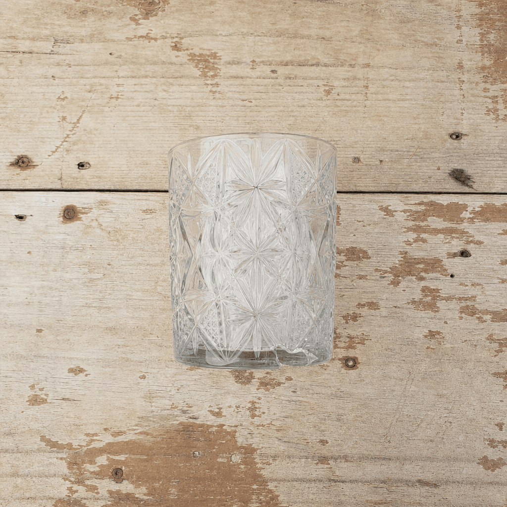 A single pressed-glass tumbler with a chip at the base, photographed straight on, on bare pine boards.