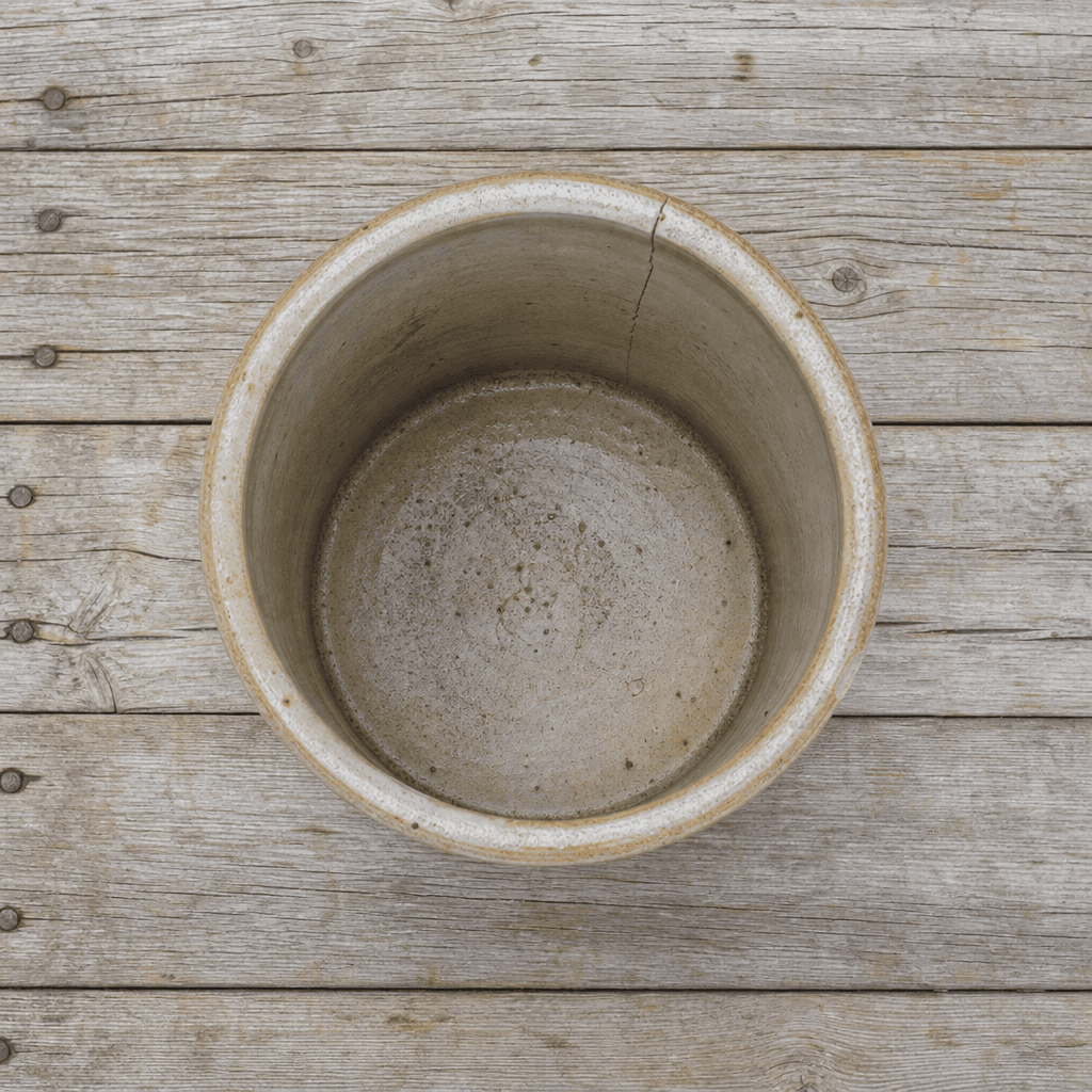 A small salt-glazed stoneware crock with a hairline crack, photographed straight on, on bare pine boards.